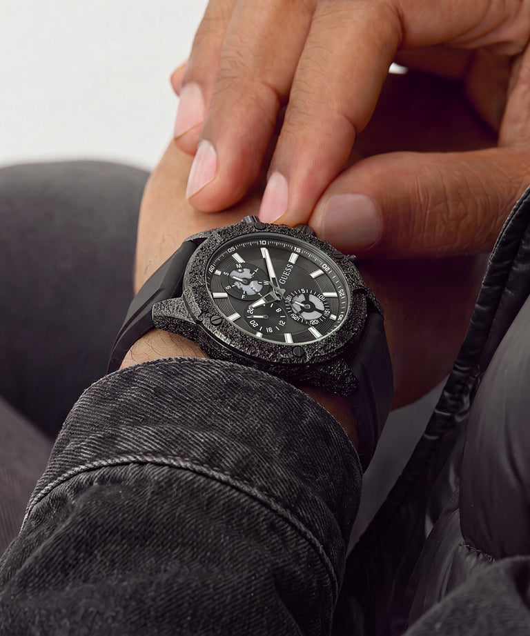 A close-up lifestyle shot of a black chronograph watch with a textured, stone-like bezel and a black rubber strap, worn on a person's wrist with their hand resting near it.