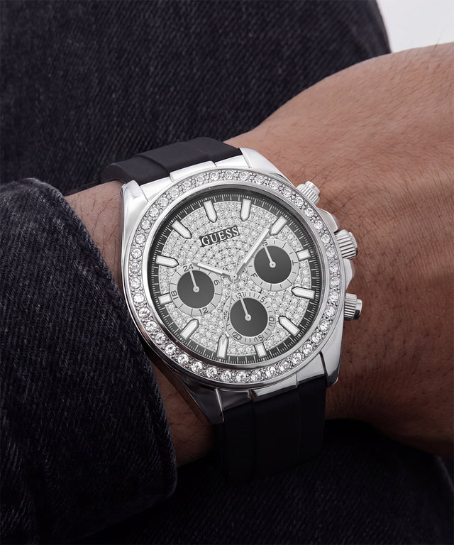 A close-up of a Guess wristwatch with a silver-tone case, diamond-encrusted bezel and dial, black subdials, and a black rubber strap worn on a person's wrist.
