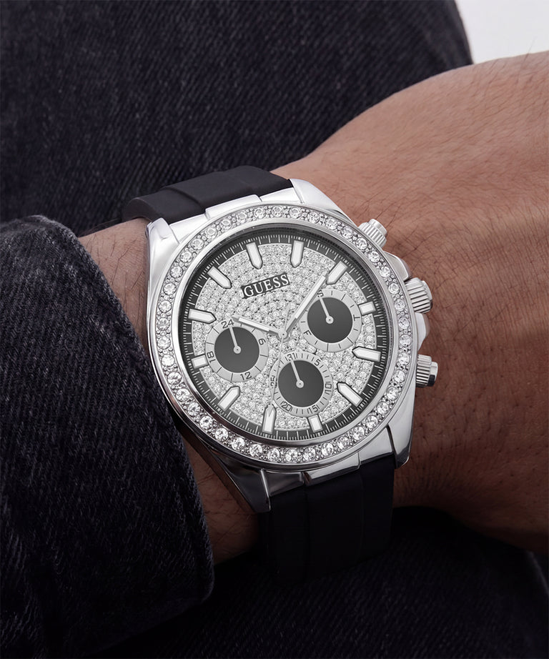 A close-up of a Guess wristwatch with a silver-tone case, diamond-encrusted bezel and dial, black subdials, and a black rubber strap worn on a person's wrist.