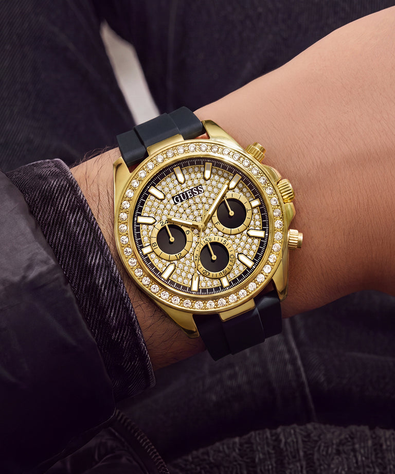 A gold-tone Guess chronograph watch with a diamond-encrusted bezel and black dial, worn on a wrist with dark denim clothing in the background.