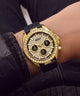 A gold-tone Guess chronograph watch with a diamond-encrusted bezel and black dial, worn on a wrist with dark denim clothing in the background.