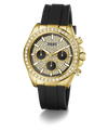 A luxury wristwatch by Guess featuring a gold-tone case encrusted with clear crystals, a black dial with three subdials, and a black silicone strap, shown against a plain white background.