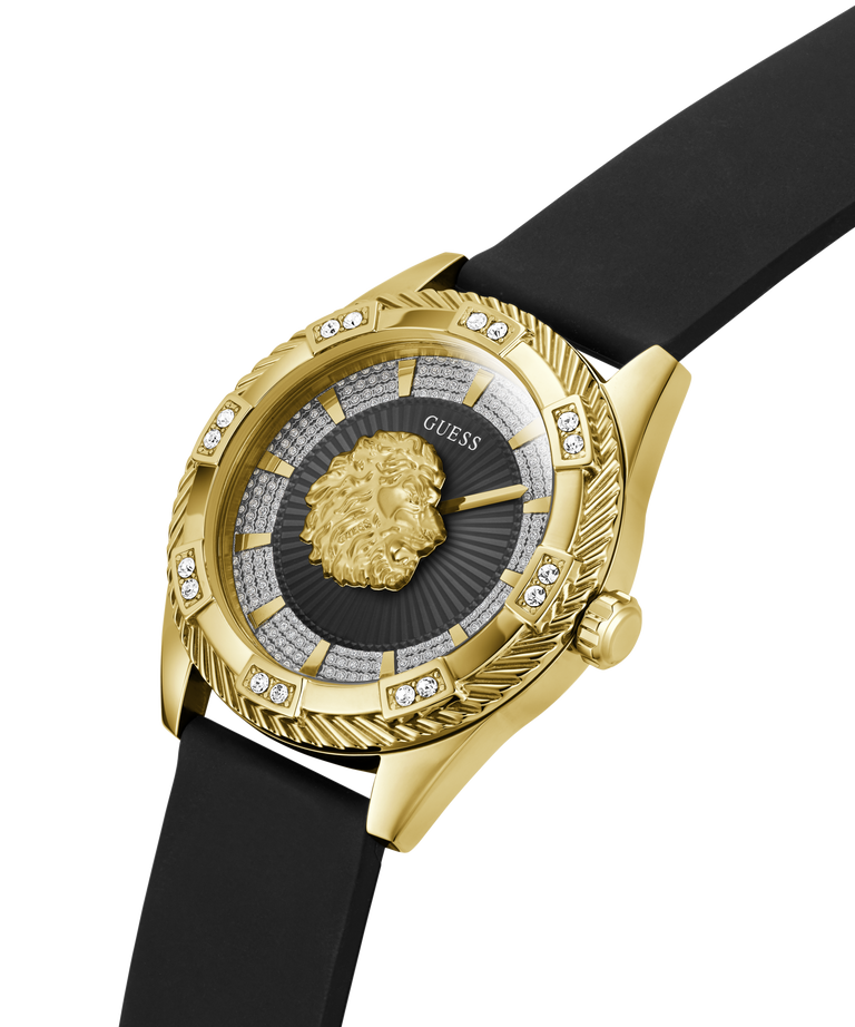 A luxury wristwatch by Guess featuring a gold-tone case with a textured bezel, crystal accents, and a central gold lion head on a dark sunburst dial. The watch is fitted with a black rubber strap and photographed on a clean white background. lifestyle