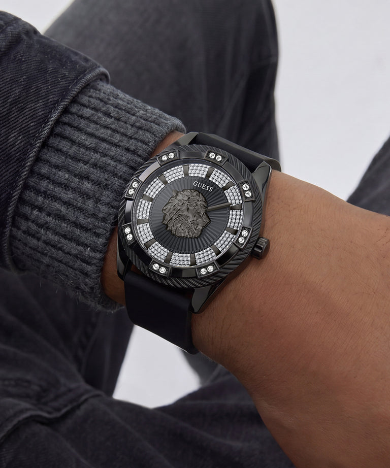 A close-up of a wrist wearing a black Versace watch with a textured dial, silver crystal-embellished bezel, and black strap, paired with a gray ribbed sweater and dark jeans.