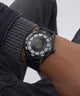 A close-up of a wrist wearing a black Versace watch with a textured dial, silver crystal-embellished bezel, and black strap, paired with a gray ribbed sweater and dark jeans.