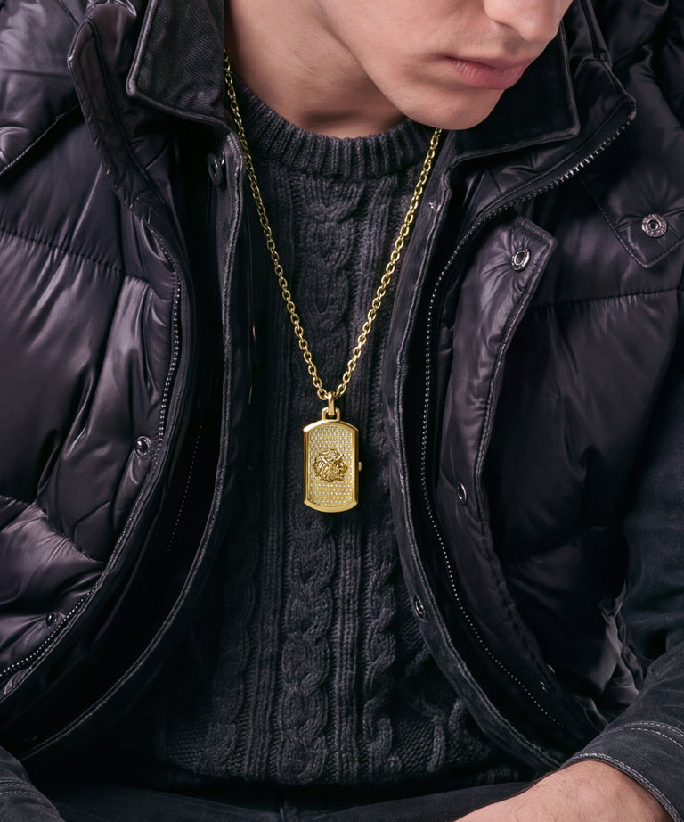A close-up of a person wearing a gold-tone dog tag pendant necklace over a black cable-knit sweater and black puffer jacket.