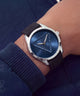 A wristwatch with a silver-toned case, a dark blue sunburst dial featuring a raised triangular pyramid design, and a black textured strap, shown worn on a person's wrist.