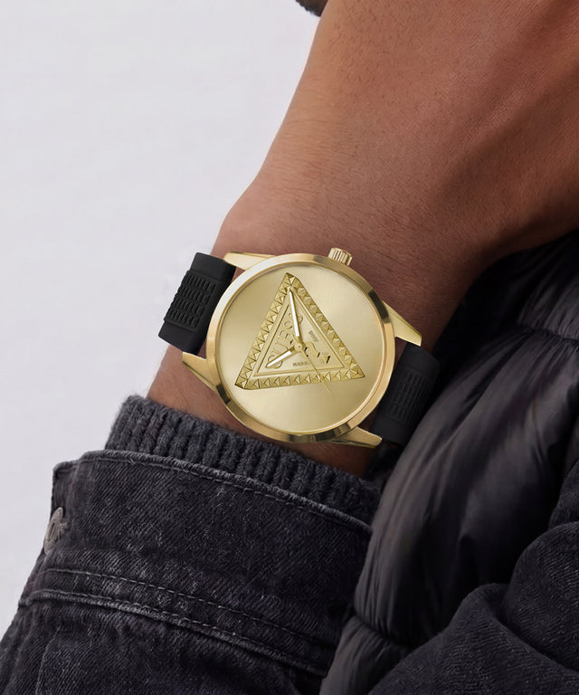 A gold-tone GUESS wristwatch with a black fabric strap, worn on a person's wrist. The watch features the brand's signature triangle logo on the dial, styled with a dark denim jacket against a light gray background.