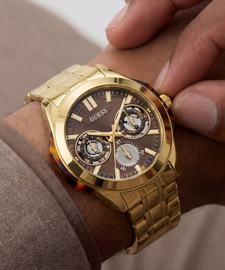 A gold-tone GUESS wristwatch with a brown dial and integrated metal link bracelet, shown worn on a person's wrist with fingers adjusting it.