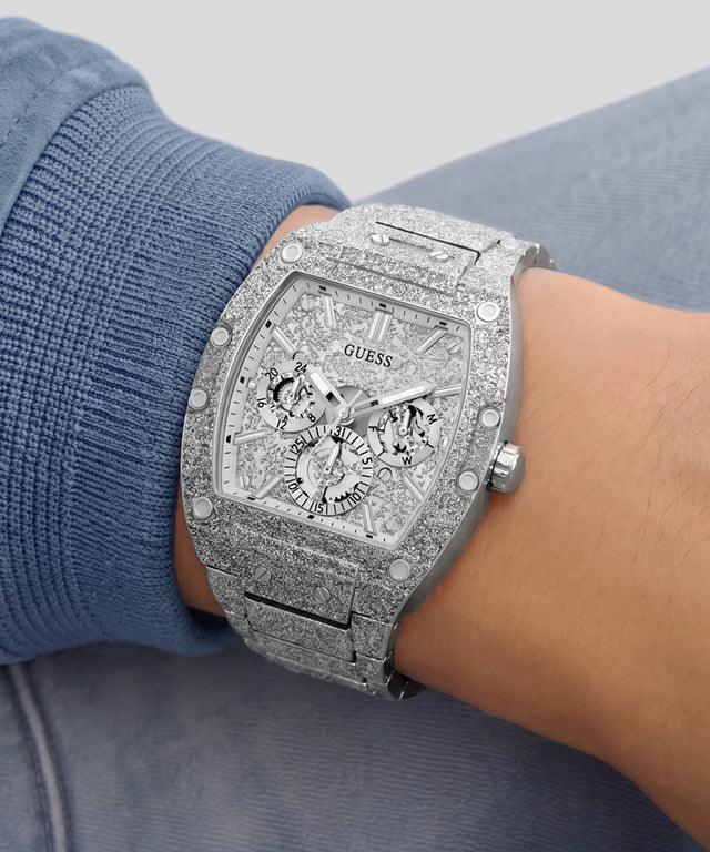 A close-up of a wrist wearing a silver-tone Guess watch with a heavily embellished crystal-look dial and matching crystal-look bracelet, paired with a blue ribbed knit sleeve.