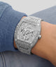 A close-up of a wrist wearing a silver-tone Guess watch with a heavily embellished crystal-look dial and matching crystal-look bracelet, paired with a blue ribbed knit sleeve.