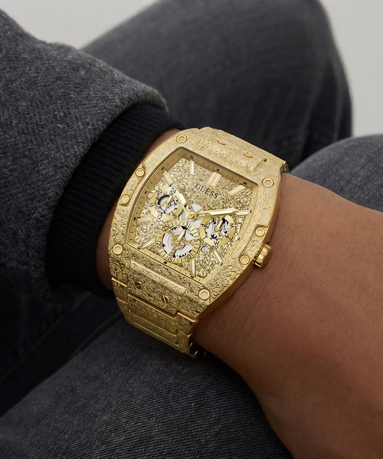 A close-up of a wrist wearing a gold-tone Guess watch with an open-heart skeleton dial and matching textured bracelet, shown against dark gray clothing.