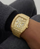 A close-up of a wrist wearing a gold-tone Guess watch with an open-heart skeleton dial and matching textured bracelet, shown against dark gray clothing.