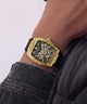 A close-up of a luxury wristwatch with a gold-tone rectangular case, skeleton dial, and black strap, worn on a person's wrist.