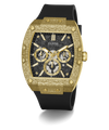 A luxury wristwatch by Guess featuring a textured gold-tone case, a black dial with three subdials, and a black rubber strap, shown on a clean white background.