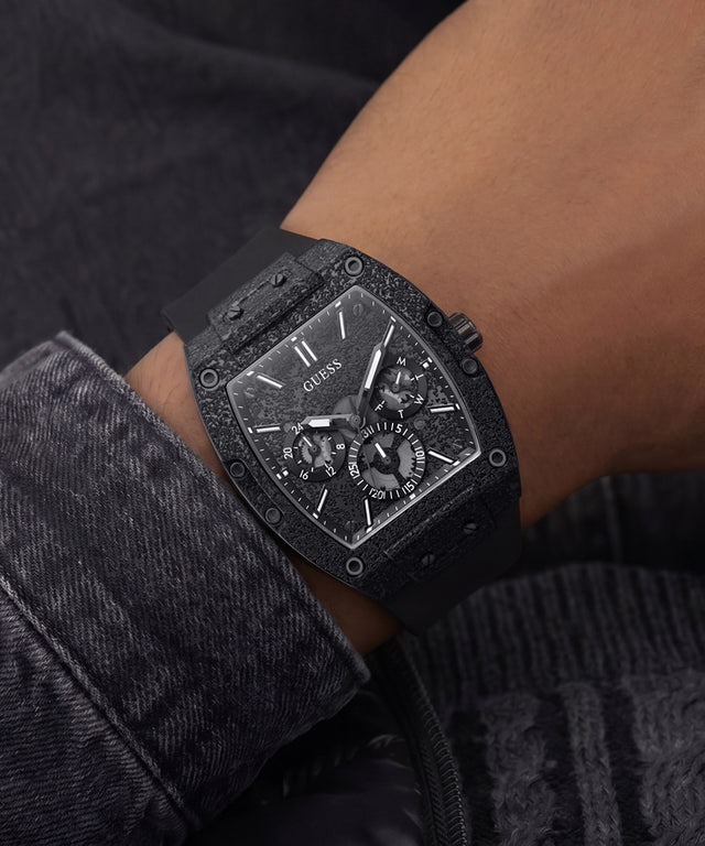 A close-up of a wrist wearing a black GUESS chronograph watch with a textured dark dial and matching black bezel, set against dark denim and skin tones.