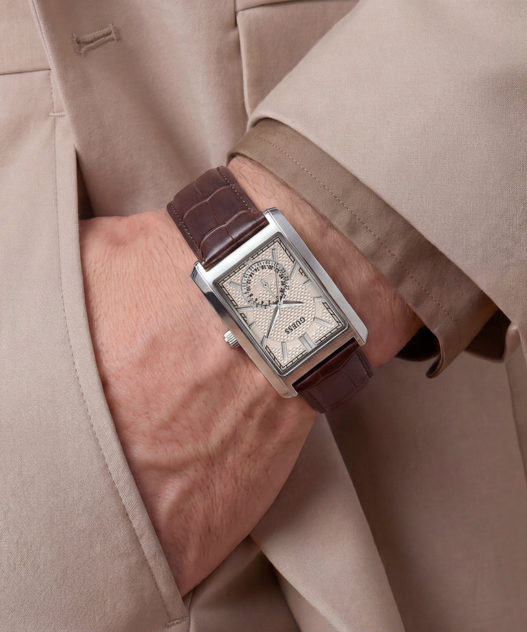 A close-up of a wrist wearing a rectangular silver-tone watch with a brown leather strap and a textured off-white dial, shown against a beige suit jacket.
