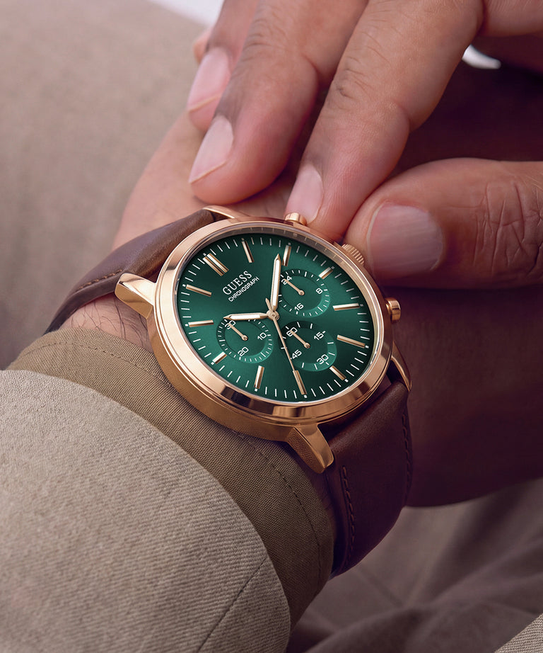 A close-up lifestyle shot of a Guess wristwatch with a green dial, rose gold case, and brown leather strap, worn on a person's wrist with fingers gently resting on the upper part of the watch.