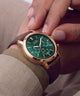 A close-up lifestyle shot of a Guess wristwatch with a green dial, rose gold case, and brown leather strap, worn on a person's wrist with fingers gently resting on the upper part of the watch.