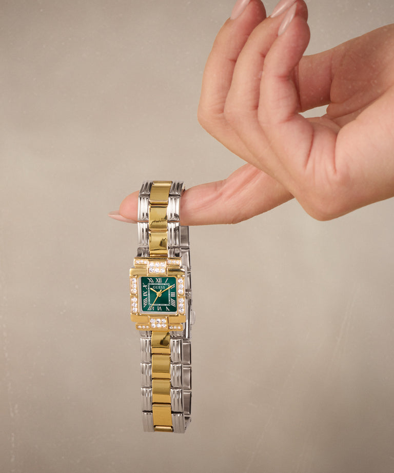 A two-tone gold and silver watch with a green dial and diamond accents, held on a person's finger against a neutral beige background.