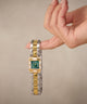 A two-tone gold and silver watch with a green dial and diamond accents, held on a person's finger against a neutral beige background.