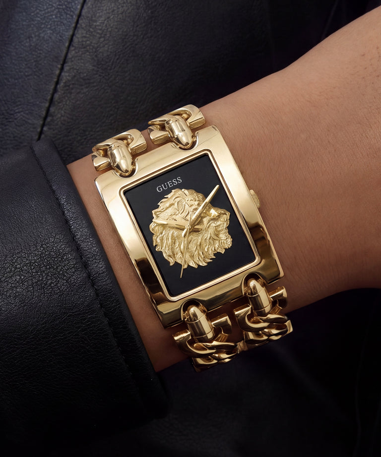 A close-up of a wrist wearing a gold-tone Guess watch with a rectangular black dial, lion head logo, and chunky chain bracelet.