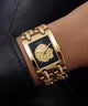 A close-up of a wrist wearing a gold-tone Guess watch with a rectangular black dial, lion head logo, and chunky chain bracelet.