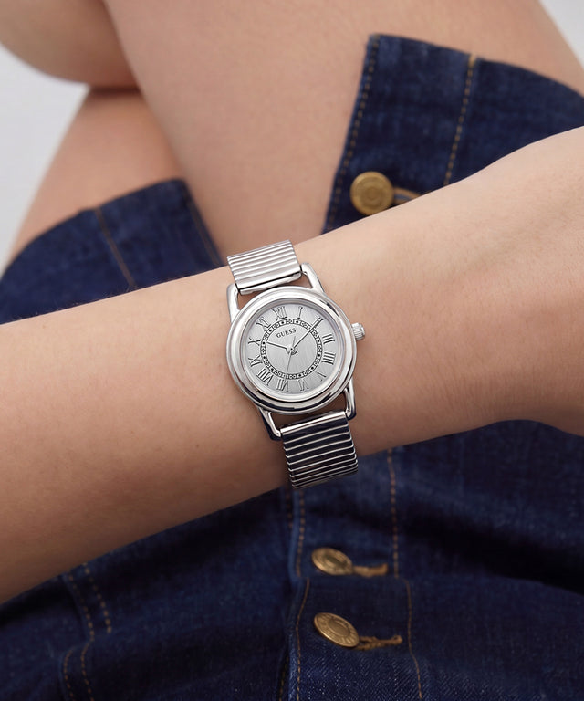 A silver-tone wristwatch with a matching metal link bracelet and a white dial, shown worn on a person's wrist. The watch has Roman numeral hour markers and a white crown, with the brand name 'GUESS' visible on the dial.