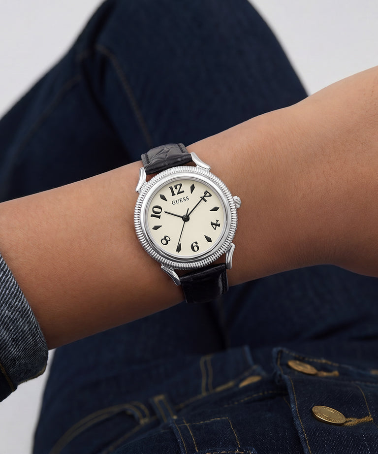 A wristwatch with a silver-tone round case, cream dial, and black leather strap worn on a person's wrist, with dark blue jeans visible in the background.