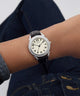 A wristwatch with a silver-tone round case, cream dial, and black leather strap worn on a person's wrist, with dark blue jeans visible in the background.