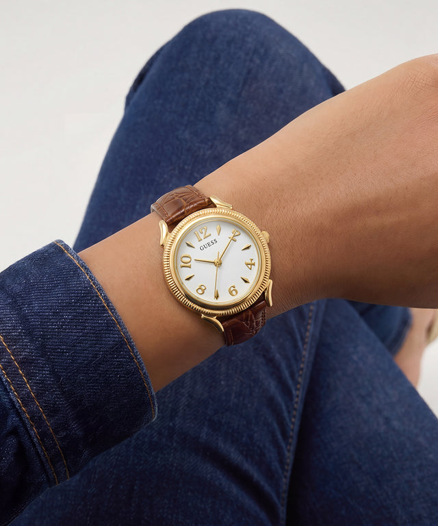 A wristwatch with a gold-tone case, white dial, and brown leather strap worn on a person's wrist, with blue denim jeans visible in the background.