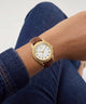 A wristwatch with a gold-tone case, white dial, and brown leather strap worn on a person's wrist, with blue denim jeans visible in the background.