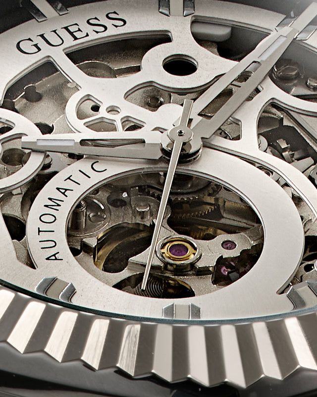 Close-up of a GUESS Automatic skeleton watch dial showing silver-toned gears, hands, and internal mechanical components through an open case.