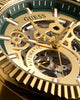 Close-up of a Guess mechanical skeleton watch with a gold-tone case, gold hands and markers, and a green dial with visible inner gears.