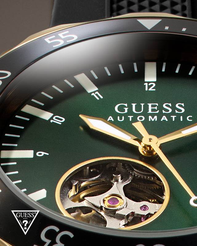 Close-up of a Guess Automatic watch with a dark green dial, black bezel, gold-tone hands and markers, and an exposed mechanical movement in the lower half.