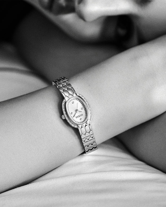 A close-up black and white photo of a woman's wrist wearing a GUESS watch with a textured metal link bracelet and an oval face.