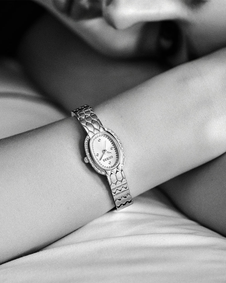A close-up black and white photo of a woman's wrist wearing a GUESS watch with a textured metal link bracelet and an oval face.
