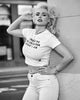 A black and white lifestyle photograph of a woman posing against a textured wall, wearing a white graphic crop top with the text 'I WANT TO BE LIKED FOR WHAT I AM' and white jeans.