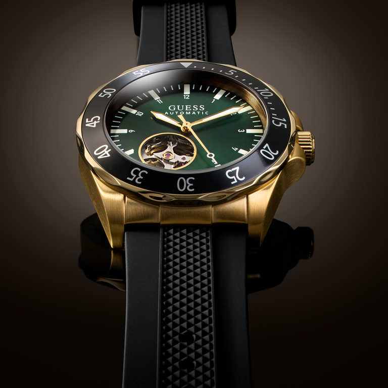 A luxury wristwatch by Guess with a gold-tone case, black bezel, green dial, and black textured strap, shown in a clean studio shot against a dark gradient background.