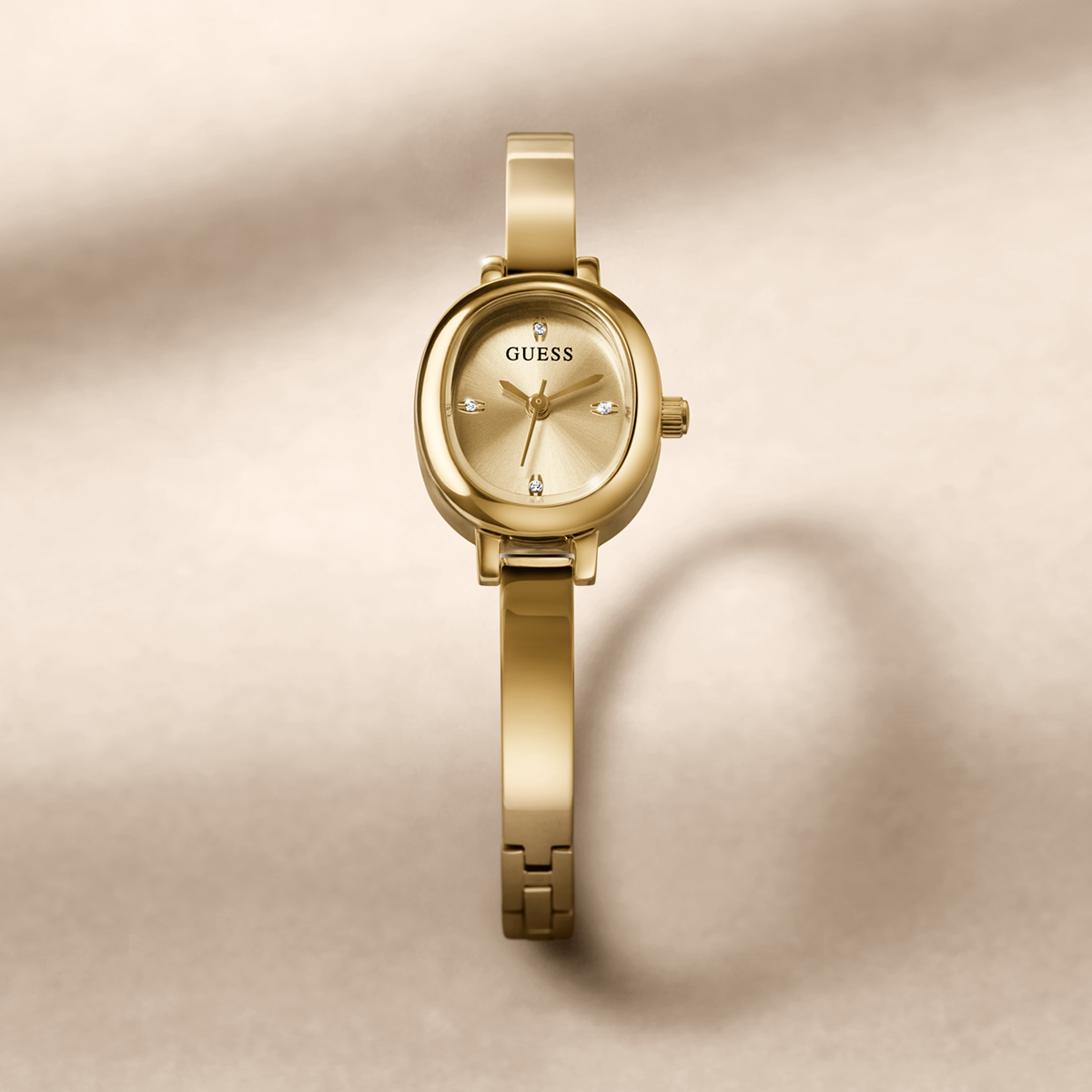 A gold-tone Guess bangle watch with a matching oval dial and integrated bracelet, centered on a warm beige background with soft shadows.