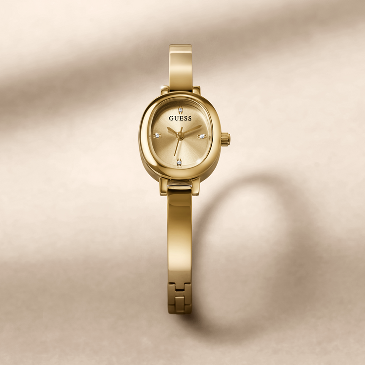 A gold-tone Guess bangle watch with a matching oval dial and integrated bracelet, centered on a warm beige background with soft shadows.