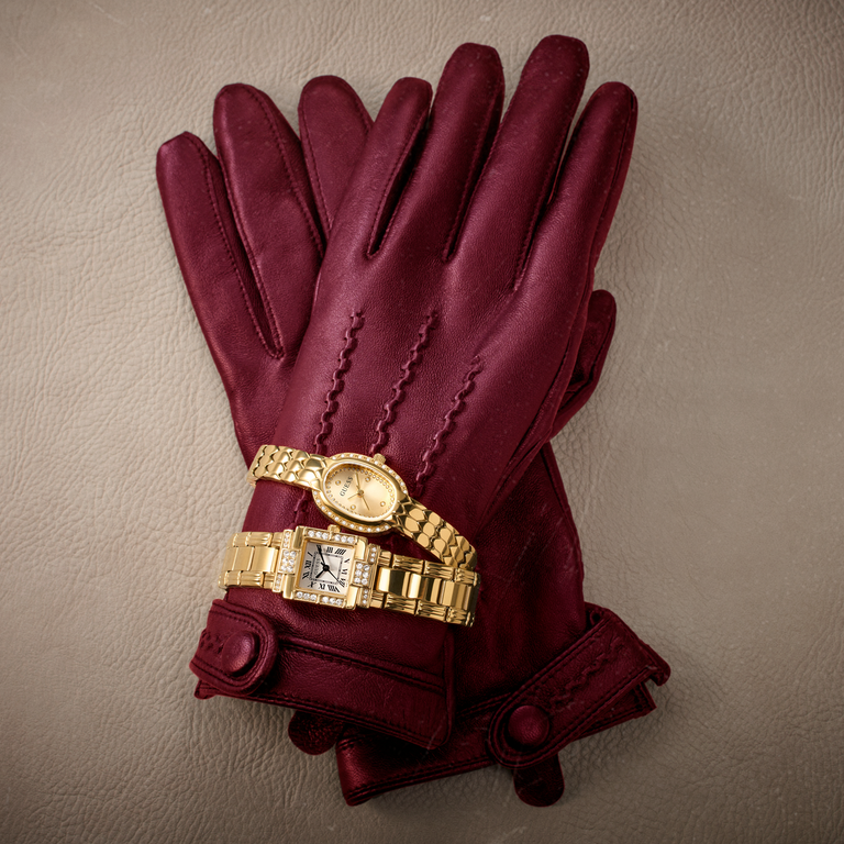 A pair of burgundy leather gloves laid flat on a textured beige surface, with two gold-tone wristwatches stacked on the back of the gloves.