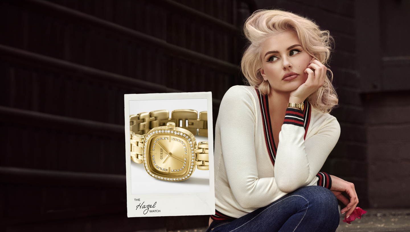 A woman in a white knit sweater and jeans sits against a dark brick wall, looking thoughtfully to the side. Overlaid on the left is a product card showing a gold-tone watch with a rectangular face and matching link bracelet.