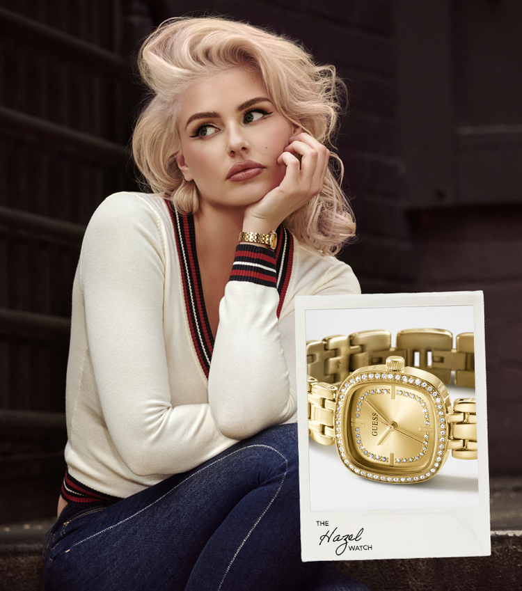 A blonde woman in a cream sweater and jeans poses next to a product insert card for a gold-tone watch with a rhinestone bezel. The insert card features the text 'The Hazel Watch'.