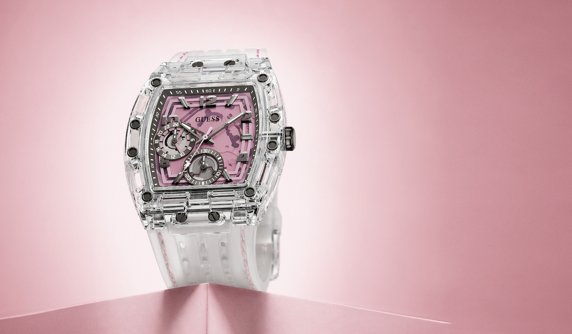 A GUESS wristwatch with a transparent clear case, a pink skeleton dial, and a white rubber strap, displayed on a pink background.