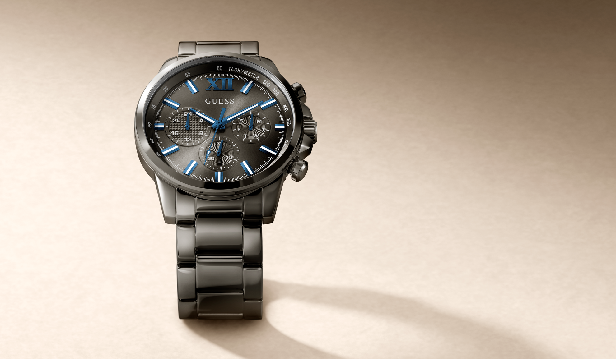 A GUESS wristwatch with a dark gray and black metal case and link bracelet, featuring a textured gray dial with blue accents, blue hands, and blue hour markers, set against a warm beige background.