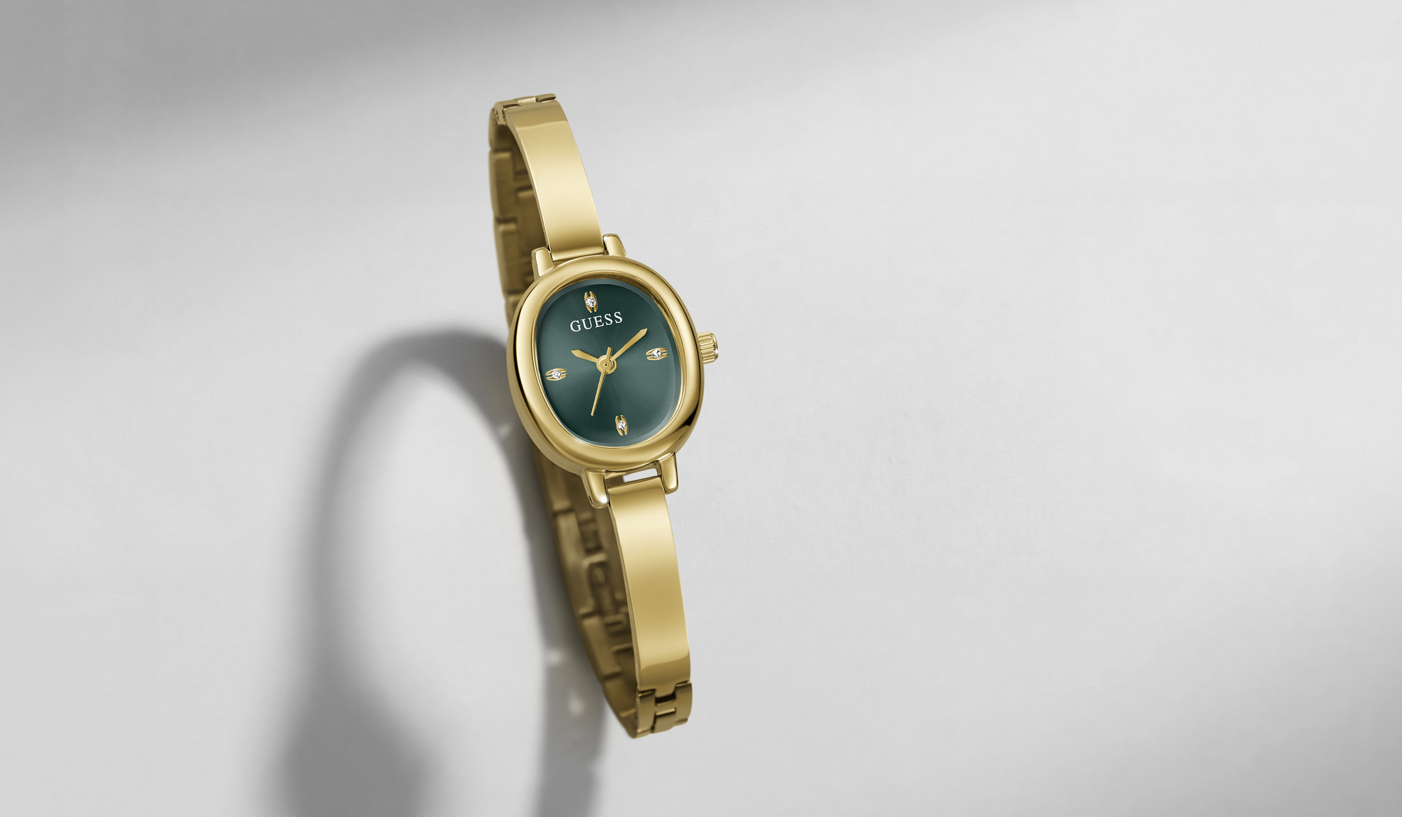 A gold-tone GUESS wristwatch with a dark green dial and matching green bezel, shown floating against a light gray background with a soft shadow.