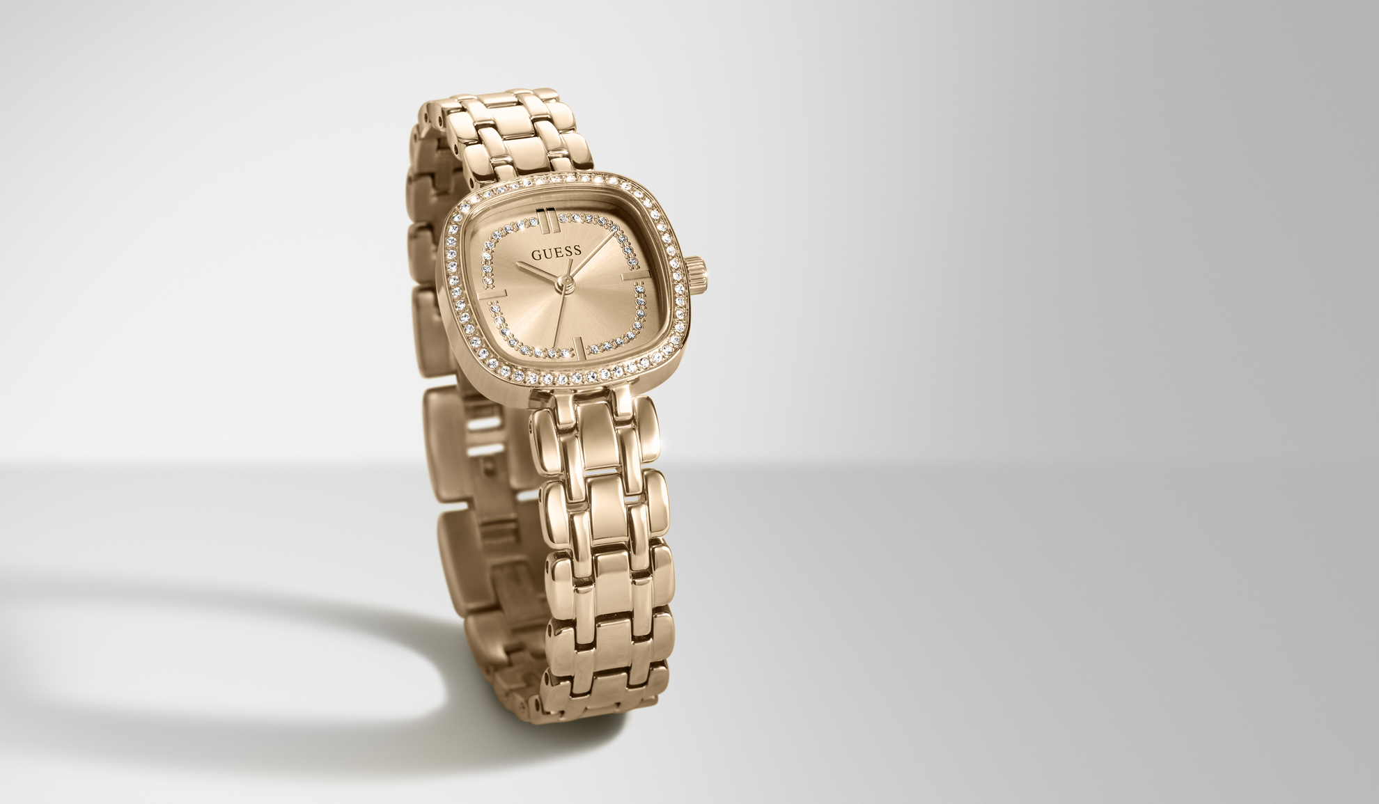 A gold-tone GUESS watch with a rectangular bezel set with small clear stones and a matching metal link bracelet, shown on a light gray studio background.