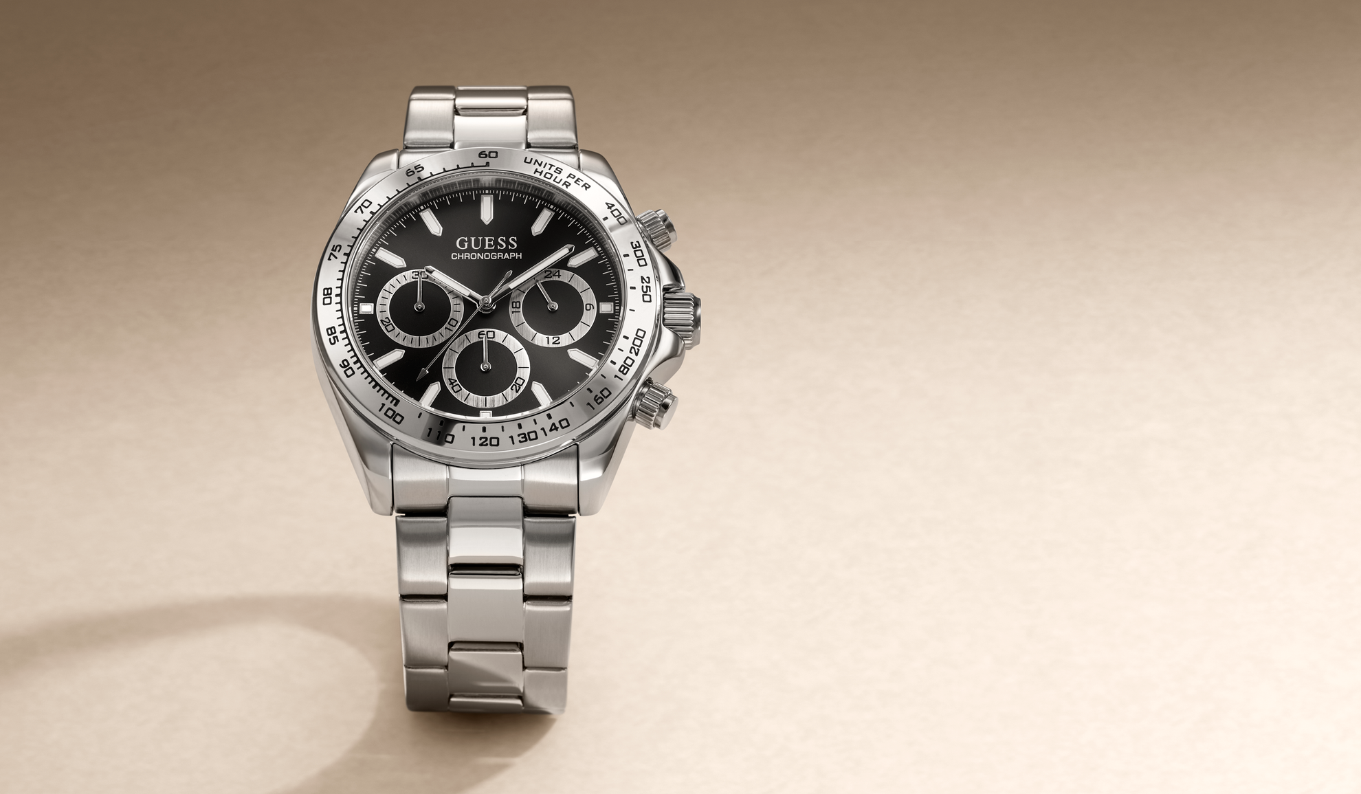 A Guess stainless steel chronograph watch with a black dial and silver link bracelet, positioned on a warm beige background.