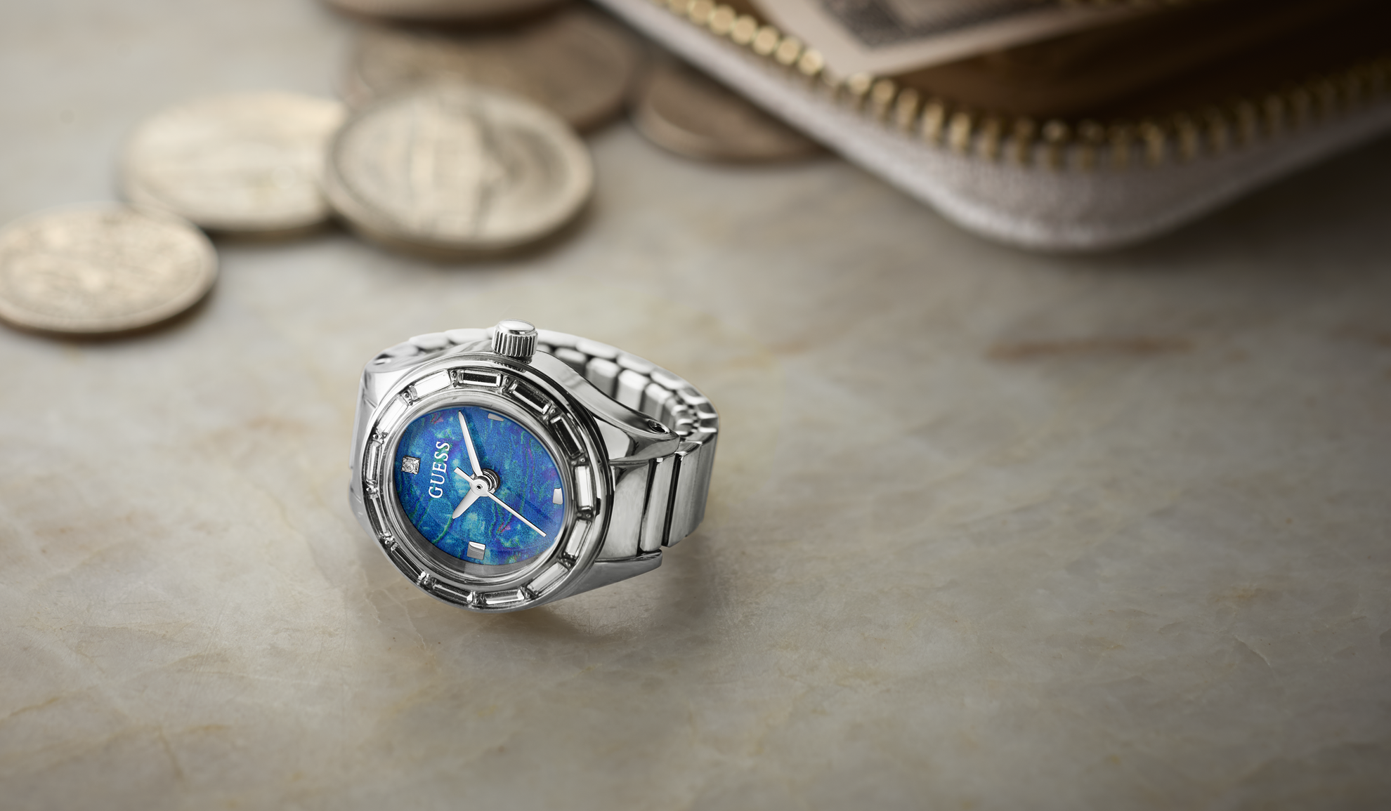A silver-tone stainless steel watch ring with a blue dial and white hands, styled on a light beige stone surface with coins and a wallet in the background.